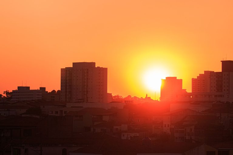 Vista urbana com casas e prédios sob o sol intenso, em um céu alaranjado no fim do dia.