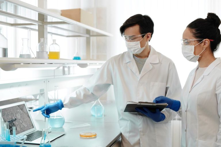 Dois pesquisadores de jaleco analisam dados em um computador dentro de um laboratório de pesquisa.
