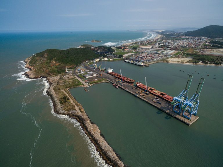 Brasil abre mercados para mamão, milho, sementes e DDG em quatro países Vista aérea do porto de Imbituba, em Santa Catarina, com cais, guindastes e navios junto à costa.