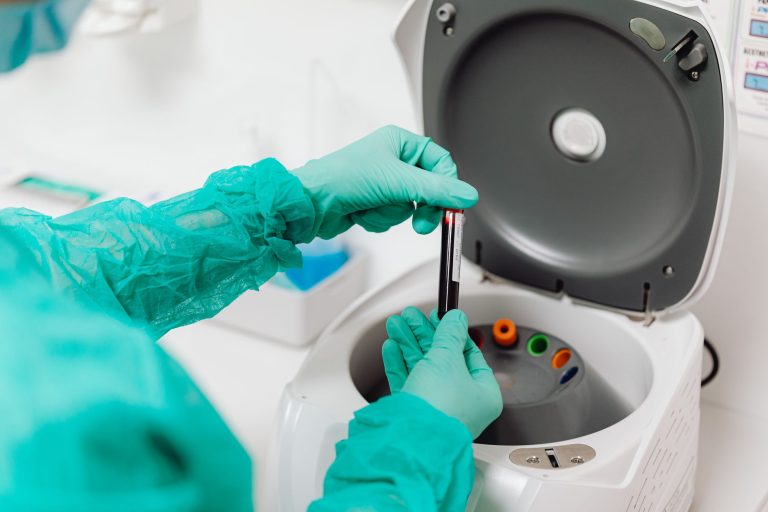 Técnica de laboratório, usando luvas, opera uma centrífuga com tubos de amostras de sangue.